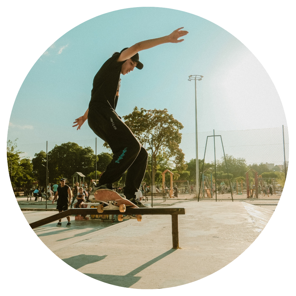 A skateboarder wearing a black t-shirt, black pants, and a black baseball cap balances on their skateboard while grinding along a metal rail in a concrete skate park under a bright, sunny sky. The image is cropped into a circle and taken from a low angle, looking up towards the skateboarder. Other park elements like trees, fencing, and possibly other people are visible in the background.