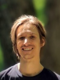 This is a headshot of a man with light brown hair that appears to be medium in length. He has fair skin and is smiling. He is wearing a dark-colored top. The background is slightly blurred and appears to be outdoors with trees visible.