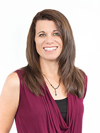 This is a headshot of a woman with long, straight brown hair and blue eyes. She has fair skin and appears to be smiling. She is wearing a sleeveless maroon top over a black top. The background is a plain white.