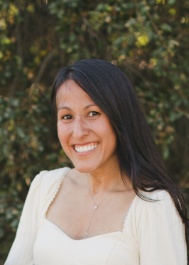 This is a headshot of a woman with long, dark hair. She has fair to medium skin, brown eyes, and appears to be smiling. She is wearing a light-colored top with a square neckline. The background is slightly blurred and appears to be outdoors with trees and foliage visible.
