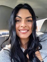 Close-up photo of a person with long, dark, wavy hair, wearing a grey top and smiling towards the camera. They appear to be inside a vehicle, with a headrest visible in the background and sunlight hitting one side of their face.