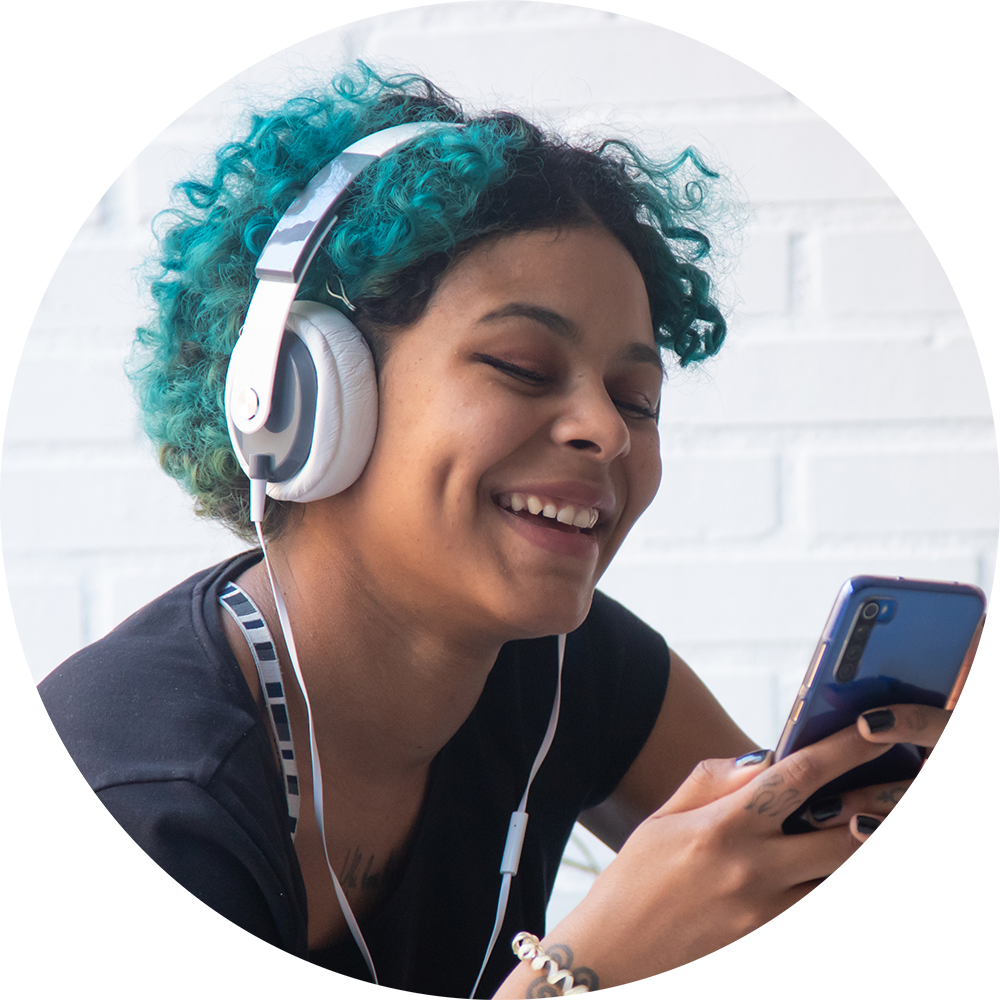 Smiling young person with teal curly hair wearing headphones and looking at a phone, sitting against a white brick wall.