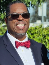 Close-up outdoor photograph of a smiling person wearing sunglasses, a dark suit jacket, a white shirt, and a red patterned bow tie. They have short dark hair and a goatee. The background is out of focus.