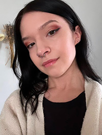 This is a close-up, selfie-style shot of a young woman with dark hair and fair skin. She has a nose ring and appears to have neutral-toned eye shadow on her eyelids. She is wearing a light-colored cardigan over a black top. The background is slightly blurred and appears to be a plain wall.