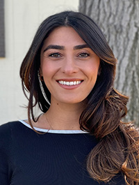 This is a headshot of a woman with long, dark, wavy hair. She has fair skin, brown eyes, and appears to be smiling. She is wearing a black top with a white neckline. The background is slightly blurred, but appears to be outdoors with a tree visible.