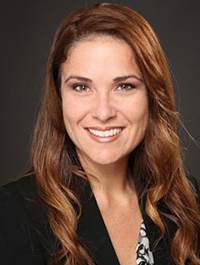 This is a headshot of a woman with long, wavy brown hair. She has fair skin, brown eyes, and appears to be smiling. She is wearing a black blazer over a patterned top. The background is a dark gray color.